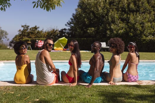 Diverse group of friends smiling and sitting at the poolside. Hanging out and relaxing outdoors in summer.