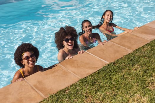 Diverse group of female friends smiling and leaning on the poolside. Hanging out and relaxing outdoors in summer.