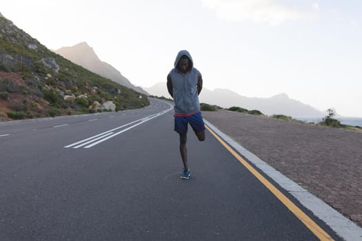 Fit african american man in sportswear stretching on a coastal road. healthy lifestyle, exercising in nature.