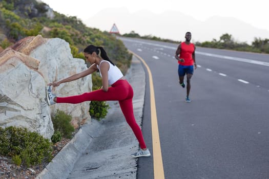 Fit african american woman in sportswear stretching while man is running on a coastal road. healthy lifestyle, exercising in nature.