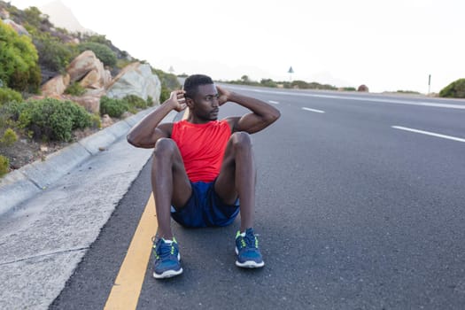 Fit african american man in sportswear doing crunches on a coastal road. healthy lifestyle, exercising in nature.