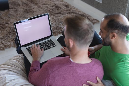 Multi ethnic gay male couple sitting on couch using laptop with copy space on screen at home. enjoying time staying at home in self isolation during quarantine lockdown.