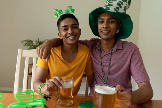 Smiling mixed race gay male couple wearing st patrick's day costumes embracing and holding beers. staying at home in isolation during quarantine lockdown.