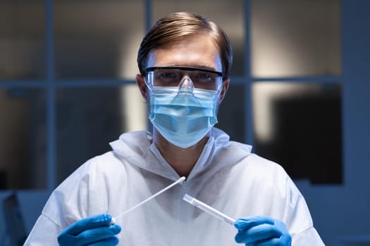 Caucasian male medical worker wearing protective clothing and face mask inspecting swab in lab. healthcare, medical research technology and hygiene during coronavirus covid 19 pandemic.