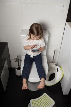 A girl dressed in a blue jeance and white t-shirt playing with a mobile phone in the bathroom.