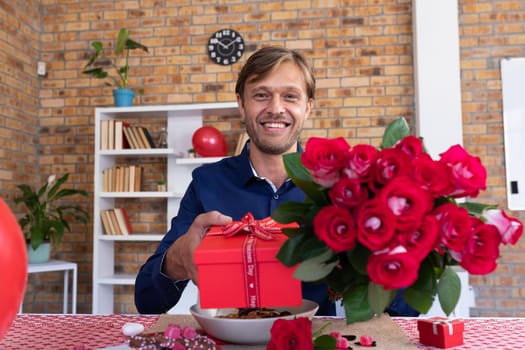 Smiling caucasian man making video call holding bunch of red roses and gift box. online communication during quarantine lockdown