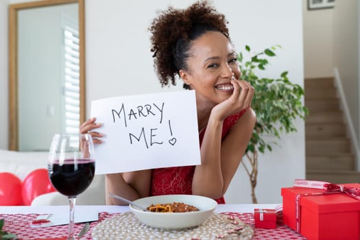 Portrait of african american woman holding placard with marry me text on videocall at home. distant celebration of valentines day online and dating concept