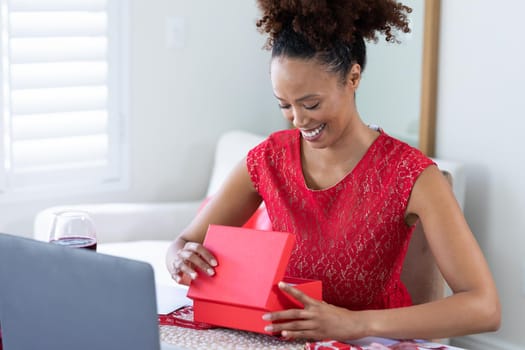 African american woman opening gift box on videocall at home. distant celebration of valentines day online and dating concept