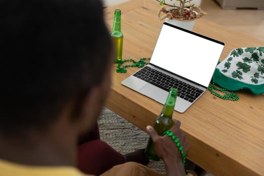 African american man with beer on st patrick's day video call using laptop with copy space on screen. staying at home in isolation during quarantine lockdown.