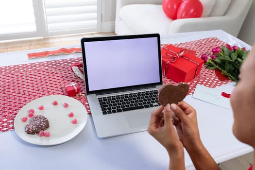 African american woman holding heart shaped cookie on videocall on laptop at home. distant celebration of valentines day online and dating concept