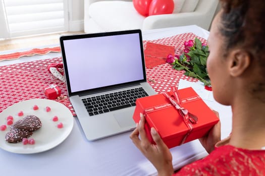 African american woman holding gift box on videocall on laptop at home. distant celebration of valentines day online and dating concept