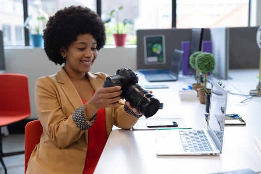 Smiling mixed race businesswoman sitting at desk using camera in creative office. social distancing in workplace during covid 19 pandemic.