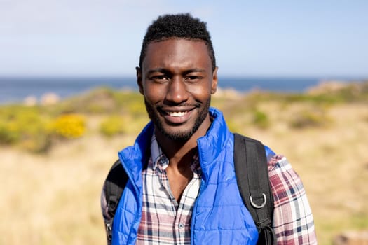 Portrait of fit african american man wearing backpack hiking on coast. looking at the camera and smiling. healthy lifestyle, exercising in nature.