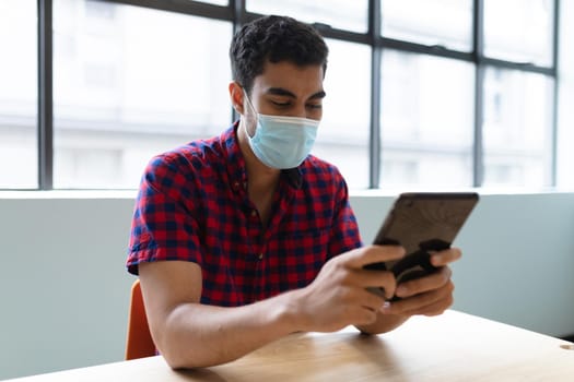 Mixed race businessman wearing face mask using tablet sitting at table. social distancing health protection hygiene in workplace during covid 19 pandemic.
