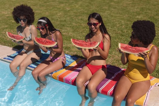 Diverse group of female friends eating watermelon sitting at the poolside. Hanging out and relaxing outdoors in summer.