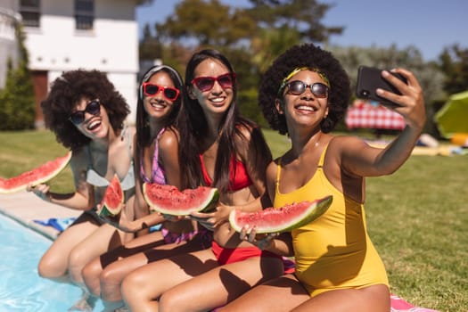 Diverse group of female friends taking selfie with watermelon sitting at the poolside. Hanging out and relaxing outdoors in summer.