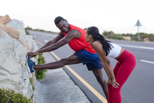 Fit african american man in sportswear stretching while woman is standing on a coastal road. healthy lifestyle, exercising in nature.