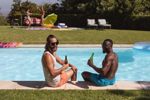 Two diverse male friends drinking beer and sitting at the poolside. Hanging out and relaxing outdoors in summer.