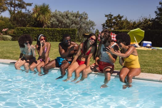 Diverse group of friends talking and sitting at the poolside. Hanging out and relaxing outdoors in summer.