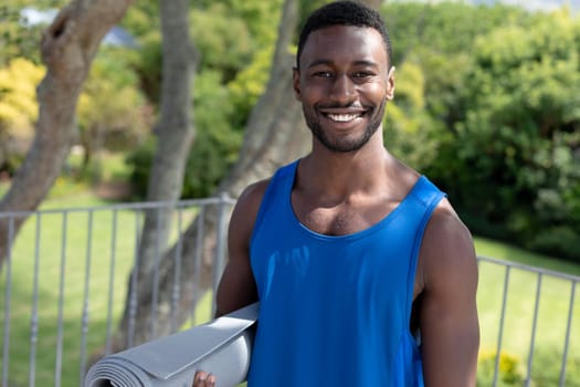 Portrait of african american man holding yoga mat smiling on sunny garden terrace. staying at home in isolation during quarantine lockdown.