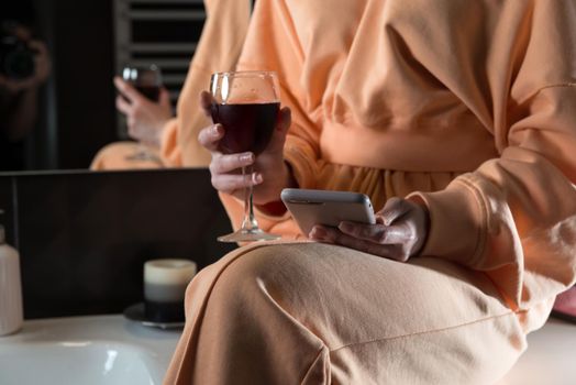 Attractive young woman in a bathroom communicating on a smartphone and drinking wine. Relaxation and leisure concept. girl wearing in orange pajamas