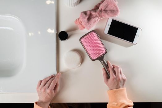 top view of bathroom accessories, toothbrush near hair brush, bow, mobile phone and face sponge on a white surface.