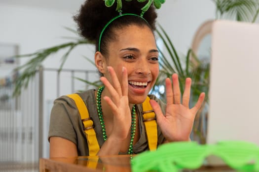 Happy mixed race woman celebrating st patrick's day making video call and waving. staying at home in isolation during quarantine lockdown.