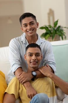 Portrait of diverse gay male couple sitting on sofa looking at camera and smiling. staying at home in isolation during quarantine lockdown.