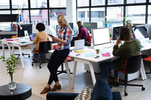 Caucasian businesswoman working in creative office. woman leaning on desk and using tablet computer, with colleagues in the background. business people and work colleagues at a busy creative office.