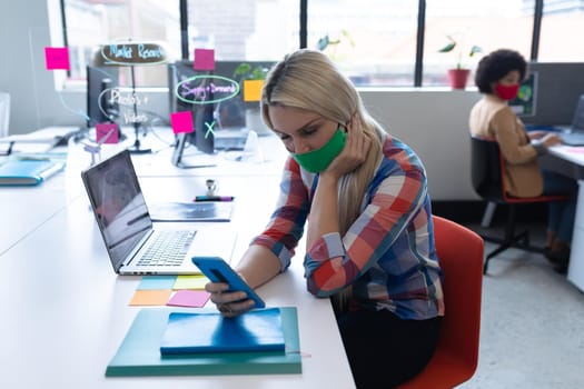 Caucasian businesswoman wearing face mask in creative office. woman sitting at desk, using smartphone. social distancing protection hygiene in workplace during covid 19 pandemic.