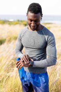 Portrait of fit african american man in sportswear using smartwatch holding water in tall grass. healthy lifestyle, exercising in nature.
