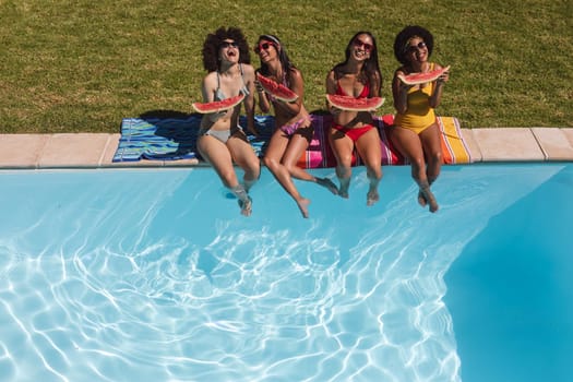 Diverse group of female friends eating watermelon sitting at the poolside talking. Hanging out and relaxing outdoors in summer.