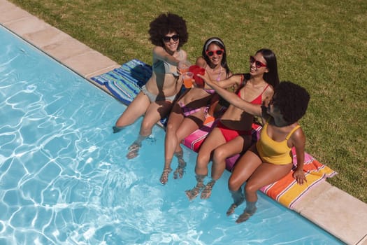 Diverse group of female friends drinking drinks sitting at the poolside. Hanging out and relaxing outdoors in summer.