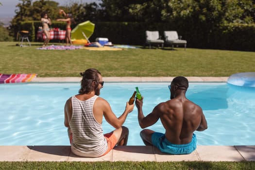 Two diverse male friends drinking beer and sitting at the poolside. Hanging out and relaxing outdoors in summer.