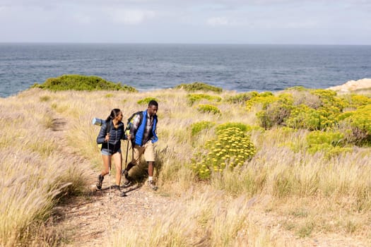 Fit african american couple wearing backpacks hiking in coastal countryside. healthy lifestyle, exercising in nature.