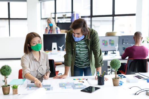 Diverse male and female colleague wearing face masks looking at blueprints and discussing. health protection hygiene in creative office during covid 19 pandemic.