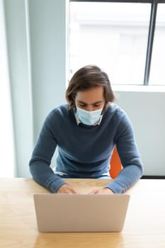 Caucasian businessman wearing face mask sitting at desk using laptop. social distancing protection hygiene in workplace during covid 19 pandemic.
