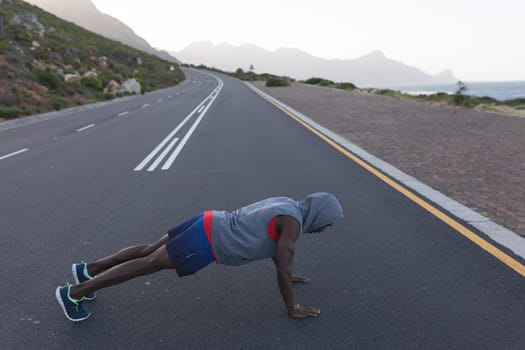 Fit african american man in sportswear doing push ups on a coastal road. healthy lifestyle, exercising in nature.