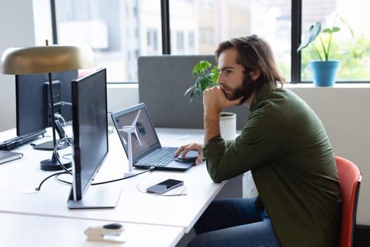 Caucasian businessman sitting at desk using laptop computer in creative office. social distancing in workplace during covid 19 pandemic.