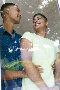 Diverse gay male couple standing by window smiling and embracing. staying at home in isolation during quarantine lockdown.