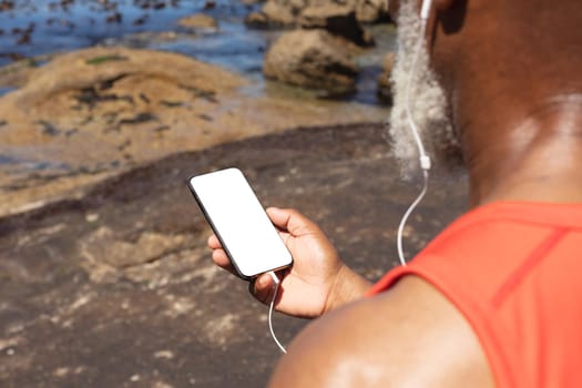 Senior african american man wearing earphones using smartphone by the sea. healthy retirement technology communication outdoor fitness lifestyle.