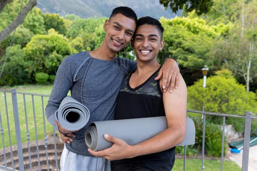 Portrait of diverse gay male couple holding yoga mats and smiling on balcony. staying at home in isolation during quarantine lockdown.