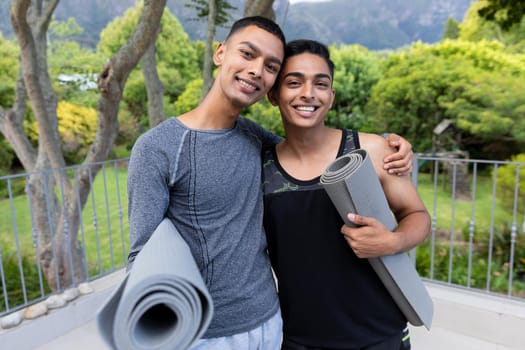 Portrait of diverse gay male couple holding yoga mats and smiling on balcony. staying at home in isolation during quarantine lockdown.