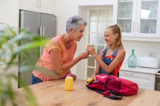 Senior grandmother giving granddaughter packed lunch and lollipop in kitchen. happy family spending time together at home.