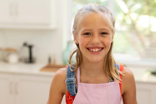 Portrait of smiling caucasian girl wearing apron standing in kitchen. staying at home in isolation during quarantine lockdown.