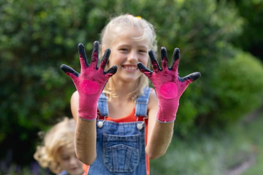 Smiling caucasian girl in garden wearing dirty pink gardening gloves, with brother in background. staying at home in isolation during quarantine lockdown.