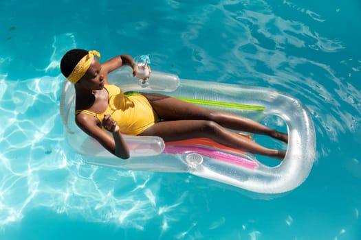 African american woman in swimming pool sunbathing on inflatable and holding drink. staying at home in isolation during quarantine lockdown.