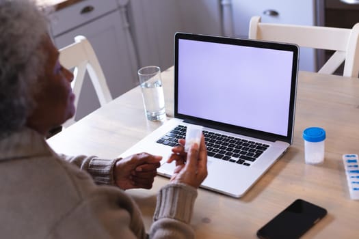 African american woman holding medication container having a video call on laptop with copy space. distant communication and telemedicine consultation concept.