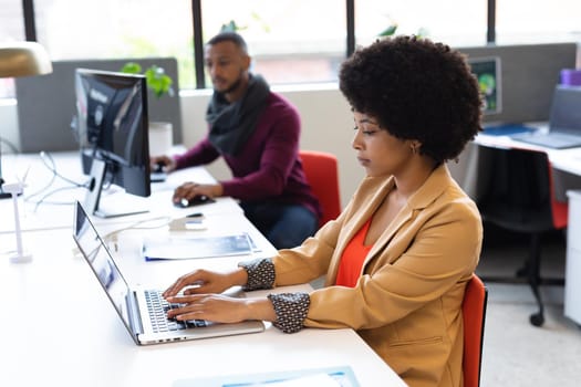 Mixed race businesswoman working in creative office. woman sitting at desk and using laptop computer, with colleague in the background. social distancing in workplace during covid 19 pandemic.