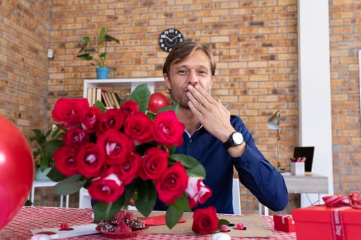 Caucasian man making video call holding bunch of red roses and blowing a kiss. online communication during quarantine lockdown